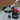 A Shop-Vac industrial vacuum cleaner in black and red is shown outdoors on a patio, with a hose connected to a black grill holding gray sand or gravel. A human hand is visible holding the hose, making this a Lifestyle image.