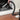 A person uses a flexible black vacuum hose to clean a fireplace. The scene shows a hand holding the hose, a black vacuum canister partially visible on the right, a white fireplace surround, and a wood floor.